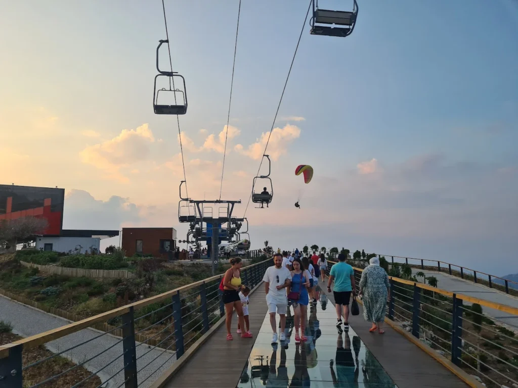 Tourists walking across a glass bridge with a chairlift passing nearby, set against a stunning mountain landscape
